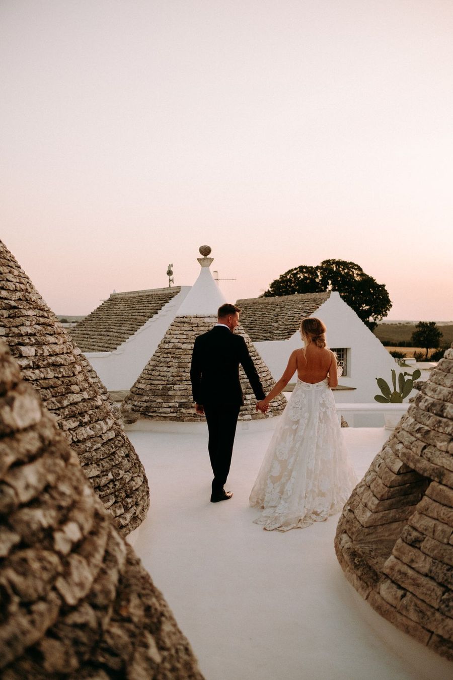 Sposi di spalle che camminano fra i tetti in pietra dei trulli, al tramonto